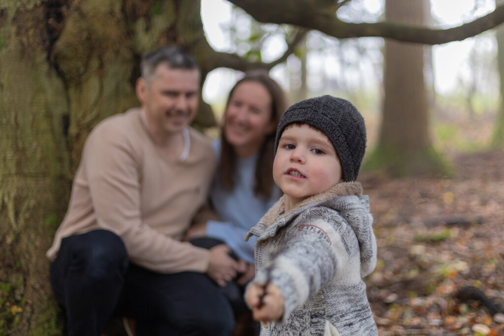 Familie sitzt vor einem Baum, Kind schaut in die Kamera, Paar unscharf im Hintergrund