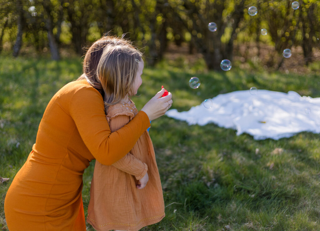 Eine Frau in einem senffarbenen Kleid umarmt ein Kind mit einem Kleid von hinten auf einer grünen Wiese. In der Luft fliegen Seifenblasen.
